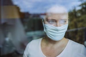 Frau schaut mit Maske während Corona-Pandemie aus dem Fenster.
