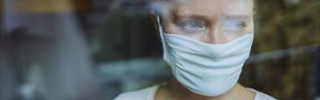 Frau schaut mit Maske während Corona-Pandemie aus dem Fenster.
